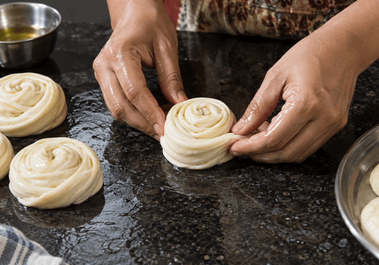 Hands shaping layered parotta dough into a spiral coil on an oiled countertop.